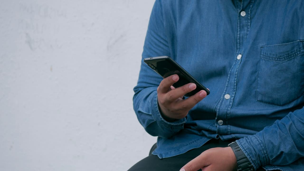 Mężczyzna w niebieskiej zapinanej na guziki koszuli przeglądający informacje za pomocą smartfona w prawej ręce