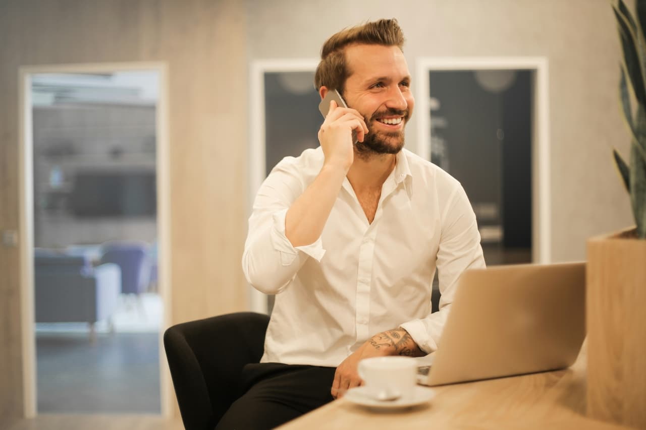 Mężczyzna rozmawiający przez telefon przy kawie przed monitorem laptopa