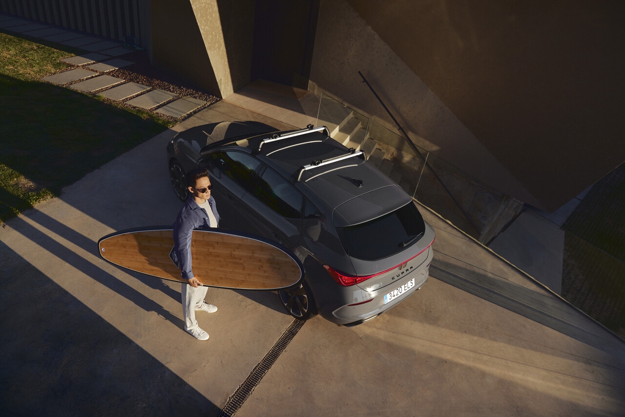 Szara CUPRA na tle minimalistycznej obudowy z mężczyzna trzymającym drewnianą deskę surfingową