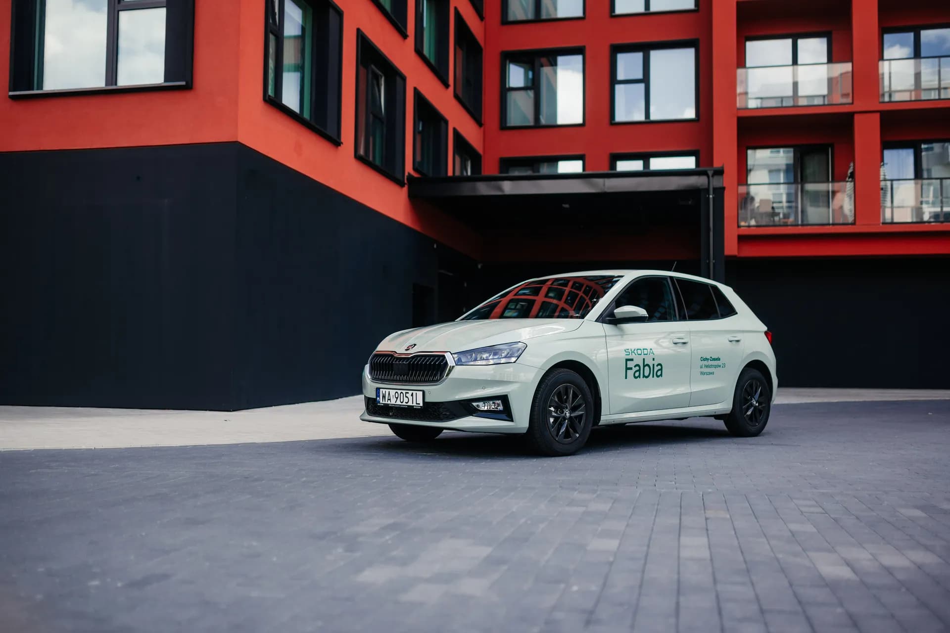 Jasnozielona Skoda Fabia przed nowoczesnym czerwonym budynkiem